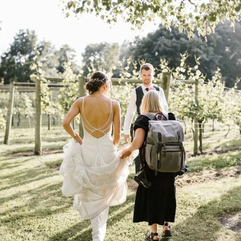 Danielle Victoria Photography Bride in a wedding dress walking through a vineyard with a person carrying a backpack.